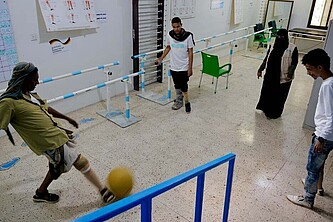 Drei Männer spielen Fußball in einem Rehazentrum. Neben ihnen steht eine Mitarbeiter von HI im Niqab.
