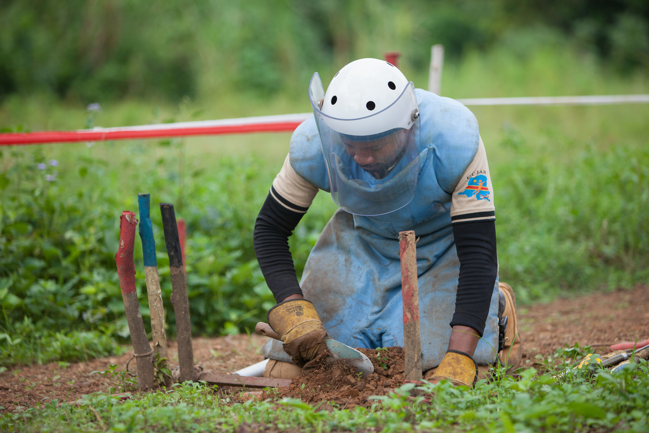 Ein Entminer kniet mit Helm und Schutzweste auf der Erde. In einem markierten Bereich sucht er mit Schaufel und Händen nach Minen.