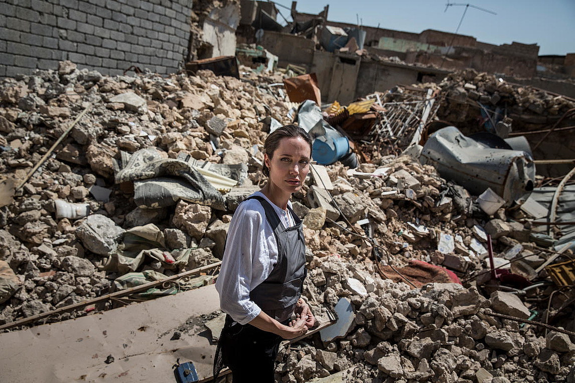 Eine Frau in Hemd und Minenschutzschürze steht inmitten von Trümmern und blickt sich besorgt nach hinten um.