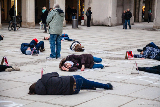 Aktivistinnen und Aktivisten liegen für einen Flashmob am Münchner Stachus auf dem Boden. Sie liegen auf oder neben aufgesprühten Leichenumrissen mit nur einem Bein. Neben ihnen stehen Warnschilder mit der Aufschrift Danger Mines.