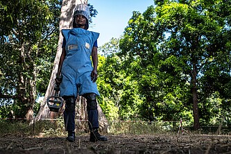 Eine Frau mit Minensuchgerät und Schutzkleidung schaut vor einem dichten Wald in die Kamera.