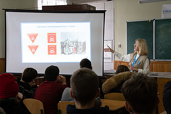 Eine Frau deutet vor einer Schulklasse auf ein Präsentation