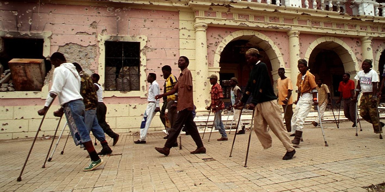 Viele Männer auf Krücken und teilweise mit fehlenden Beinen marschieren vor einer zerschossenen Häuserfront