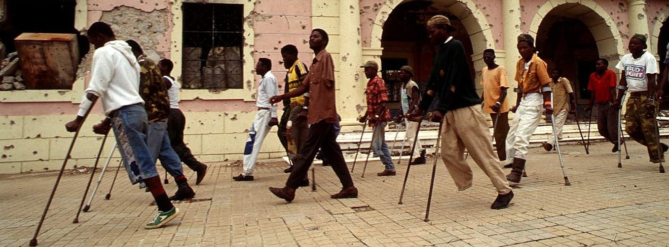 Viele Männer auf Krücken und teilweise mit fehlenden Beinen marschieren vor einer zerschossenen Häuserfront