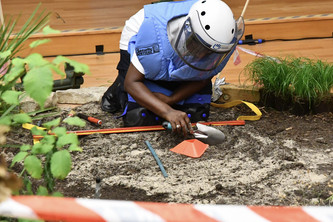 Eine Person in Schutzmontur kniet und gräbt vorsicht in einem künstlichen Minenfeld