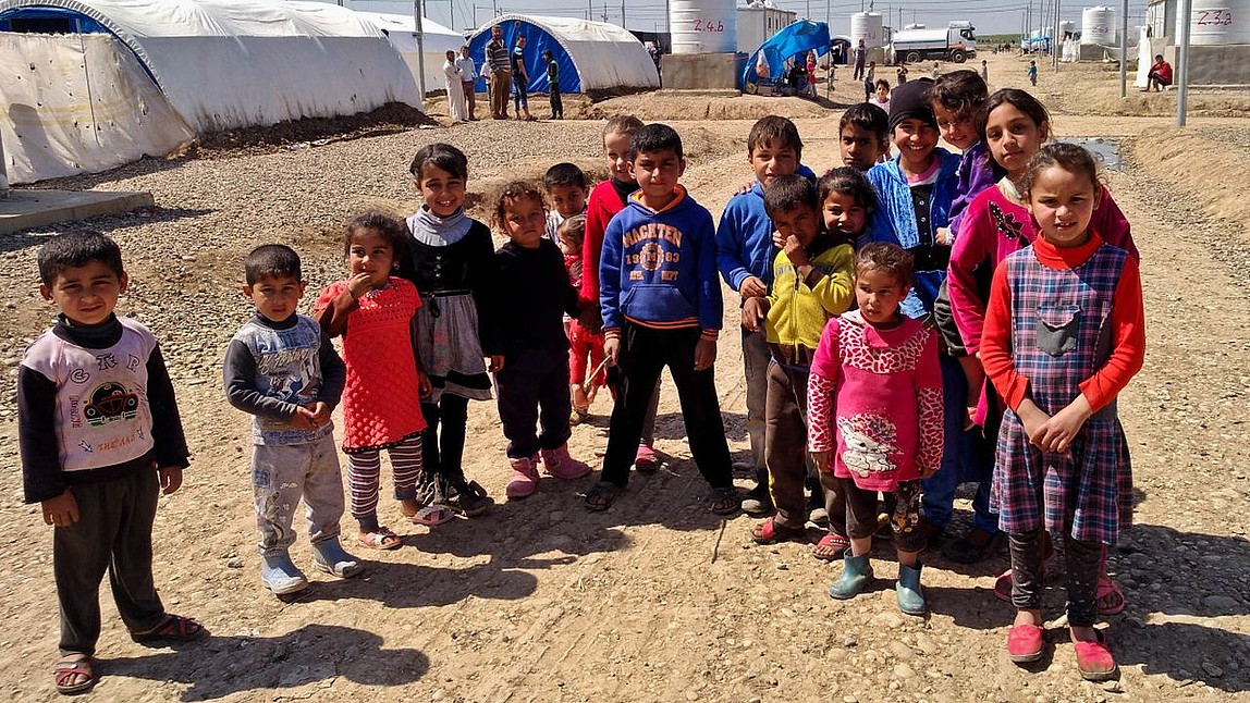 Mehrere Kinder schauen in die Kamera. Im Hintergrund ein Flüchtlingslager.