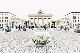 Vorne liegt das Mahnmal mit einem Kranz. Dahinter stehen Menschen in einem Halbkreis und halten sehr große Portraits hoch. Im Hintergrund das Brandenburger Tor.