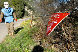 Fotografie zeigt Warnschild mit Totenkopf und Schrift "Danger Mines" an Stacheldrahtzaun, das auf Minengebiet hinweist. Im Hintergrund Schutzanzug tragende Person auf Weg neben bewachsenem Gelände erkennbar.