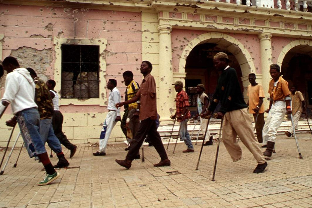 Viele Männer auf Krücken und teilweise mit fehlenden Beinen marschieren vor einer zerschossenen Häuserfront